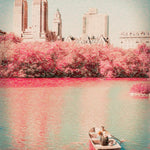 Photograph of a lone rowboat gliding across reflective water, framed by flowering trees and distant city architecture, conveying calm, nostalgia, and quiet romance.